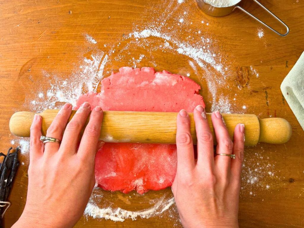 A woman is rolling out pink dough with a wooden rolling pin.