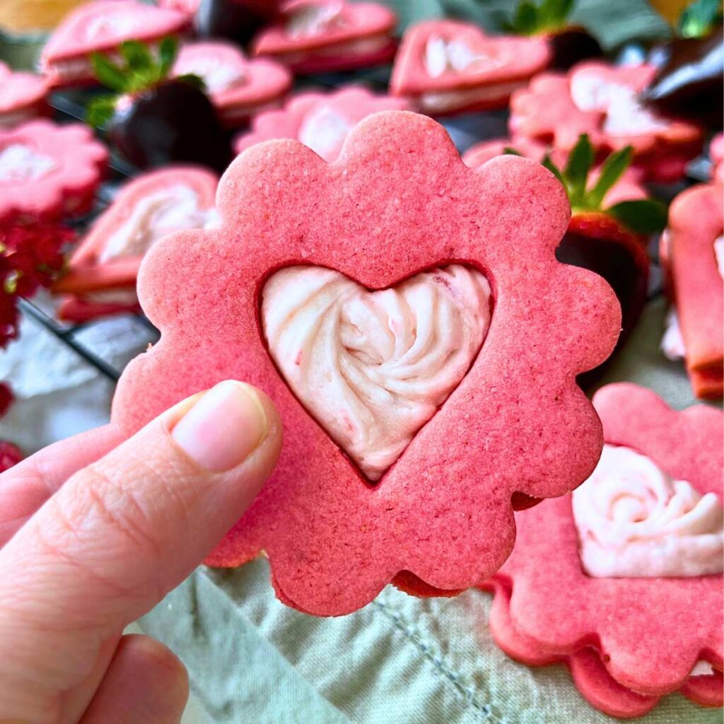 A woman is holding a strawberry sandwich cookie with a heart cut out in the middle.