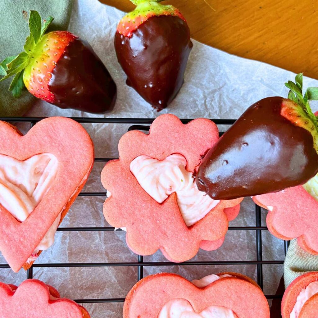 Strawberry sandwich cookies with heart cutouts. There are chocolate dipped strawberries .