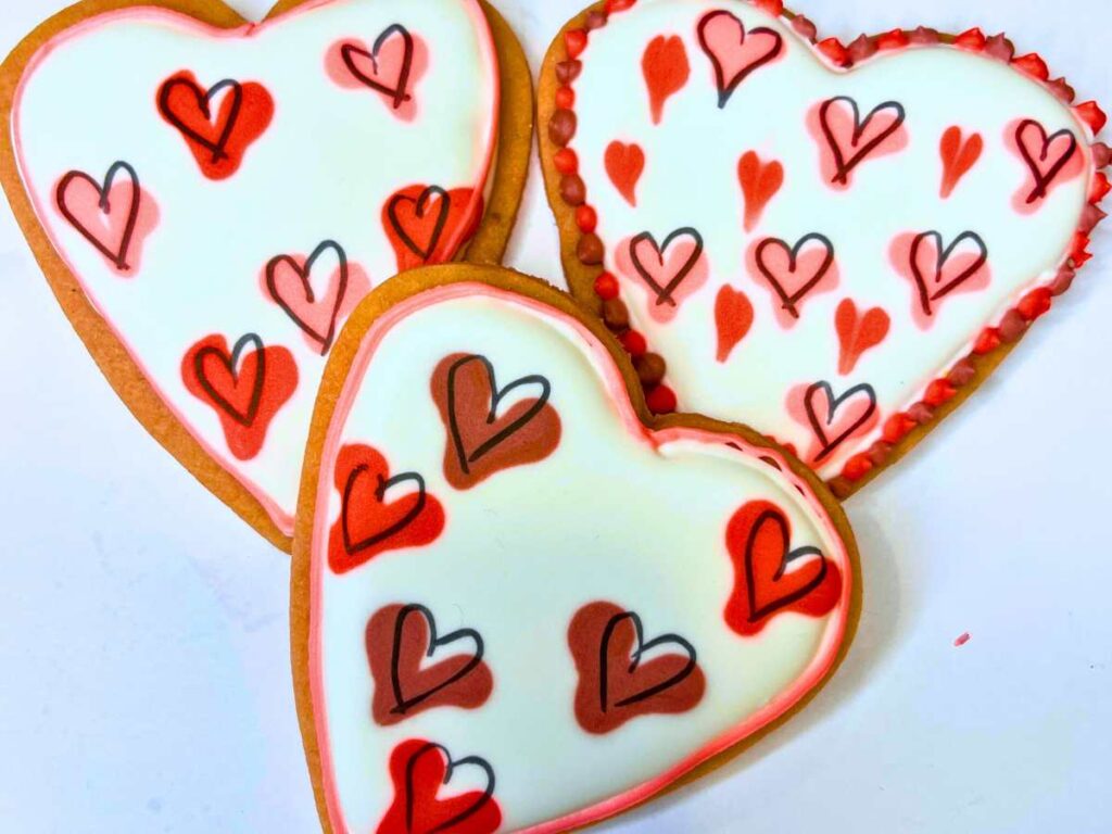 Three pink cookies with heart decorations in royal icing.