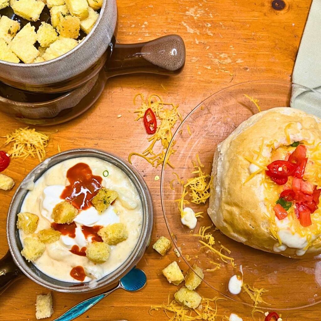 Potato soup in a bowl bowl and another in a bread bowl. There are croutons, cheese and sour cream on top.