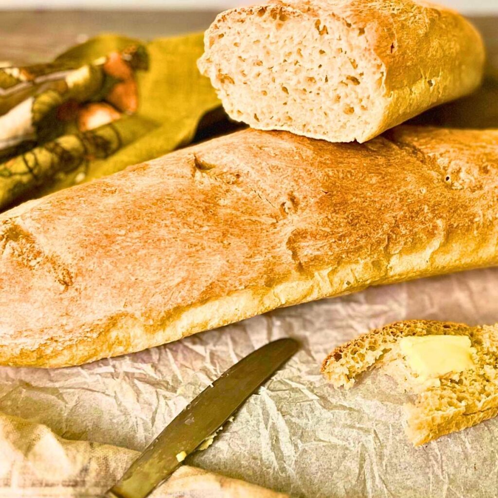 A loaf of French bread on a wooden table. There is a partial sliced loaf on top.