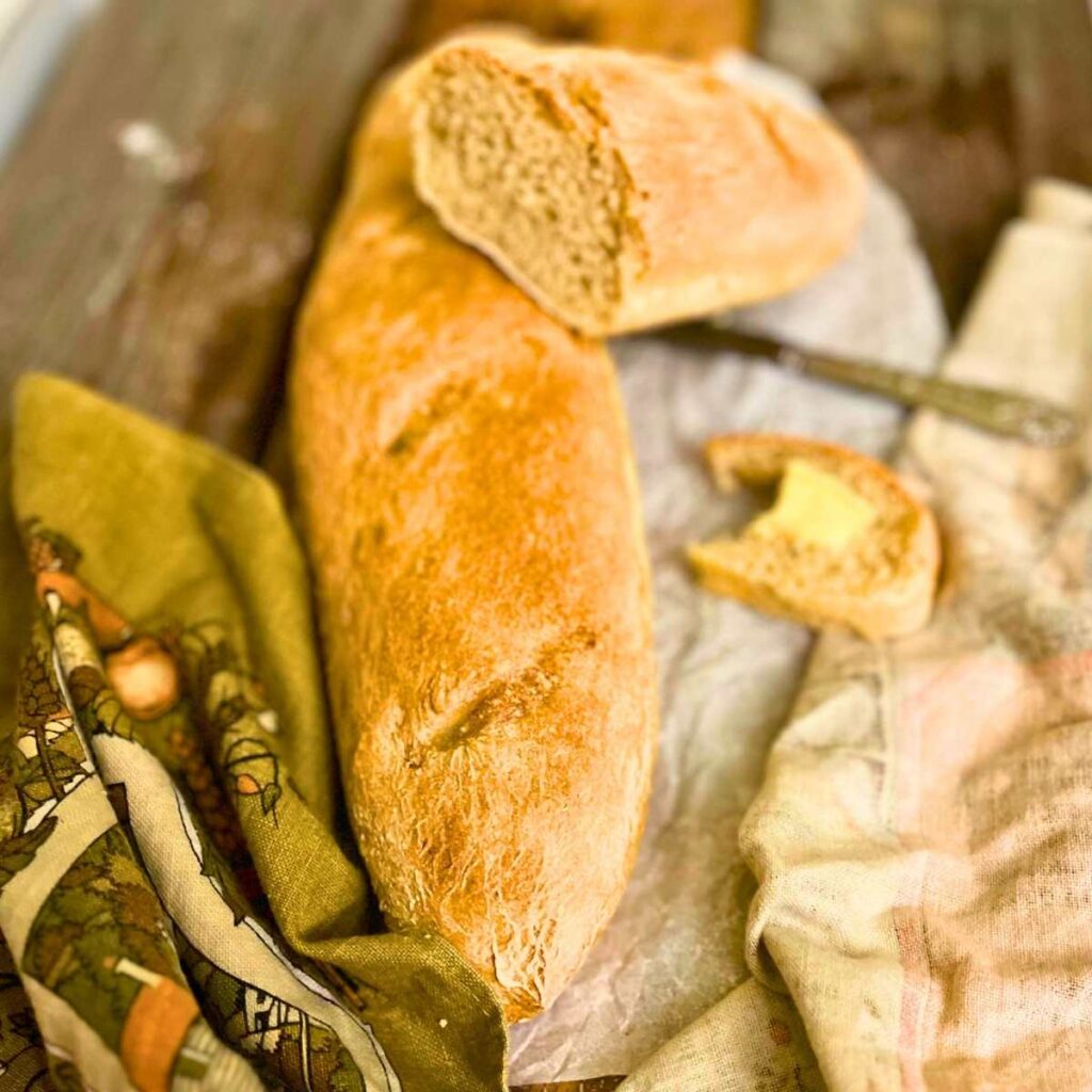 A baguette on a wooden table. There is another baguette on top that is sliced in half.