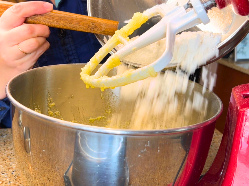 Woman adding a dry flour mixture to the bowl of a stand mixer with the paddle attachment.