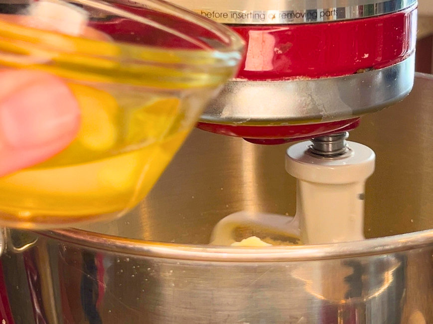 Woman adding an egg in a small glass bowl to the bowl of a stand mixer with the paddle attachment.