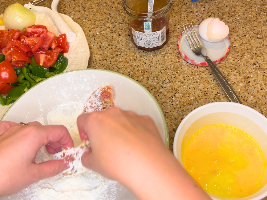 A woman coating a piece of pork in a cornstarch mixture. There is a bowl with a beater egg beside.