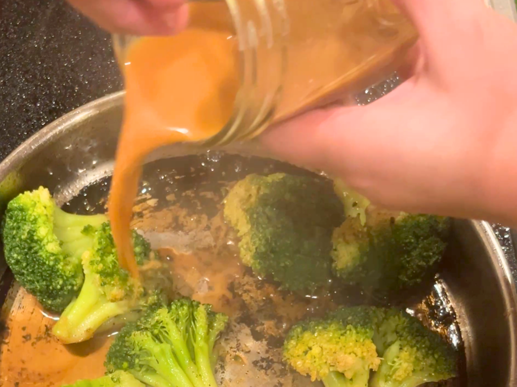 Woman pouring a brown liquid from a canning jar into a frying pan with broccoli in it.