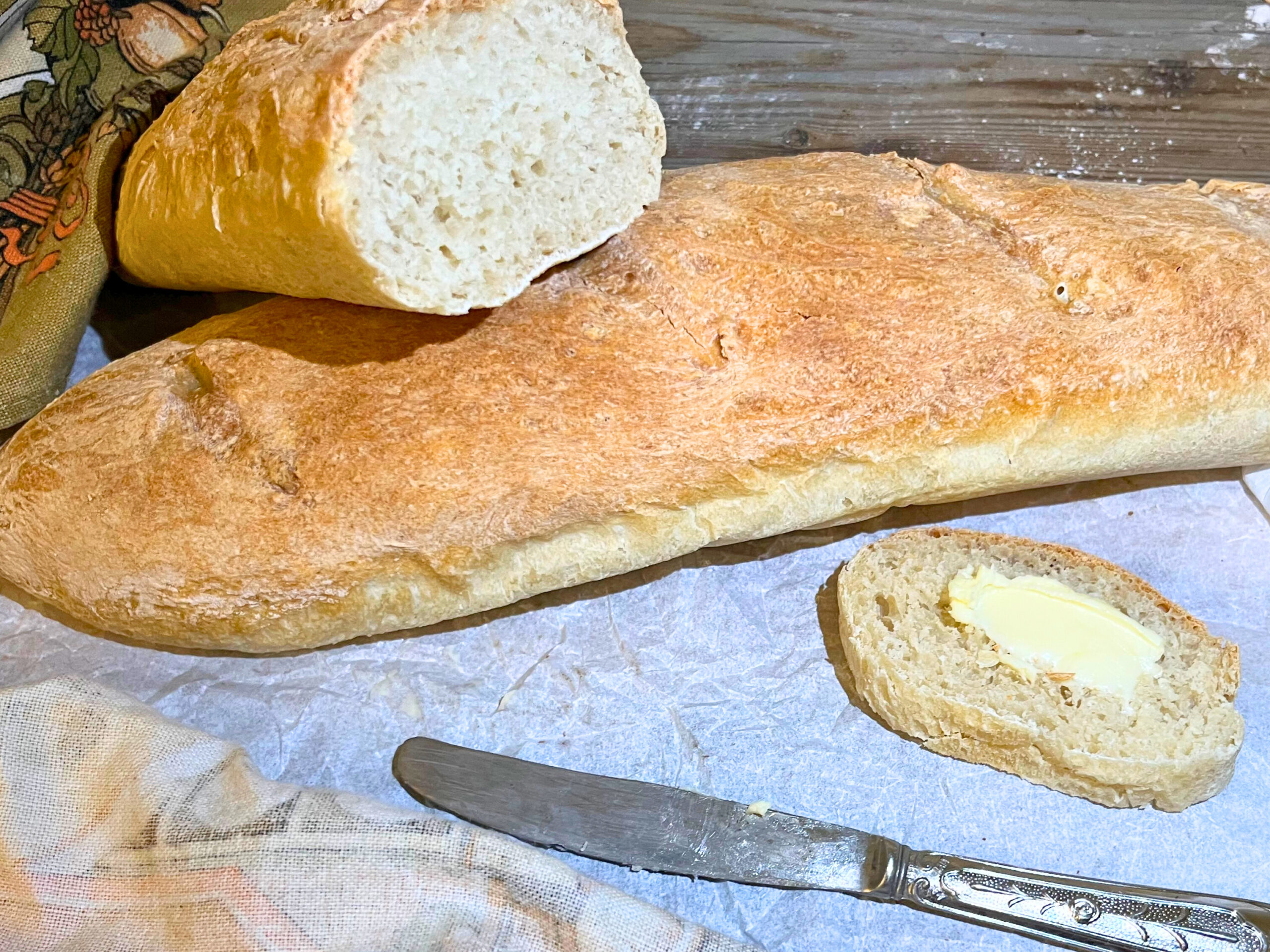 A baguette on a piece of parchment paper. A loaf cut in half and a slice of bread in the foreground with butter on top.