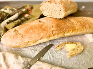 A French baguette on a piece of parchment paper there is a cut baguette and a slice with butter in the foreground.