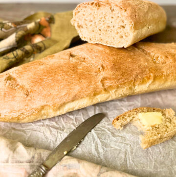 A French baguette on a piece of parchment paper there is a cut baguette and a slice with butter in the foreground.