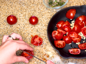 Woman cutting tomatoes in half. There is a frying pan with tomatoes and garlic in it.