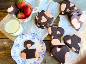 Two tone cookies, chocolate and pink. There is a glass of milk and a cup of strawberries on the table as well.