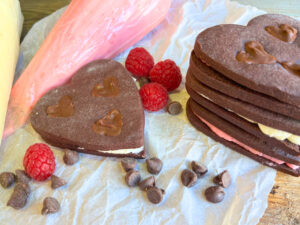 A stack of heart chocolate sandwich cookies on a piece of parchment paper. There is chocolate chips and berries and a piping bag in the background.