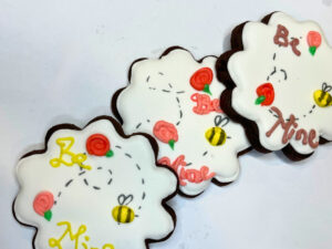 Three chocolate sugar cookies with a bee and flowers. Be mine is written on the cookies.