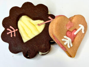 Two sandwich cookies, one is chocolate with vanilla icing, and the other is pink with pink icing. They have heart cut outs and there is an icing arrow on top.