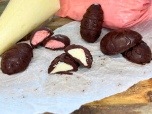 Chocolate eggs on a piece of parchment paper. Two of the chocolates are cut in half. One has a white filling and one has pink filling. Two piping bags are in the background full of filling.