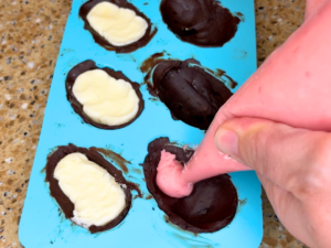 A woman filling a chocolate egg mold with pink filling.