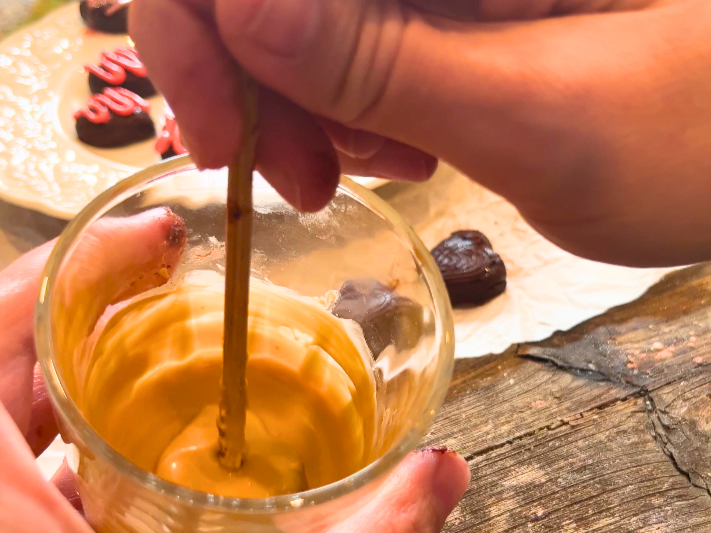 A woman using a wooden skewer to dip into a cup with melted caramel.