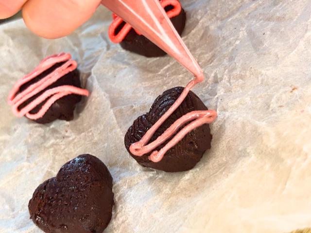 A woman using a piping bag to drizzle pink icing on a chocolate heart.