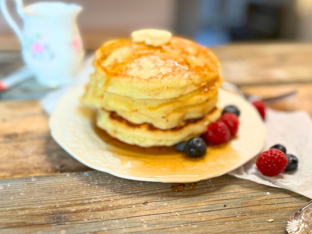 A stack of pancakes on a white plate. There is raspberries and blueberries on the plate. There is syrup and butter on top.