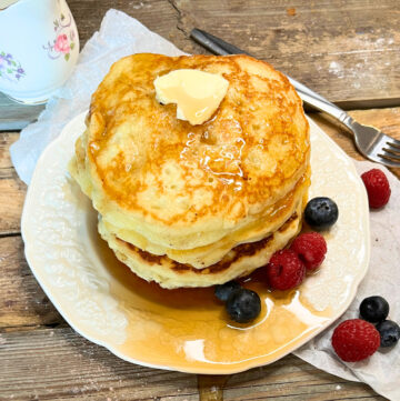 A stack of pancakes on a white plate. There are berries around and maple syrup and butter on top.