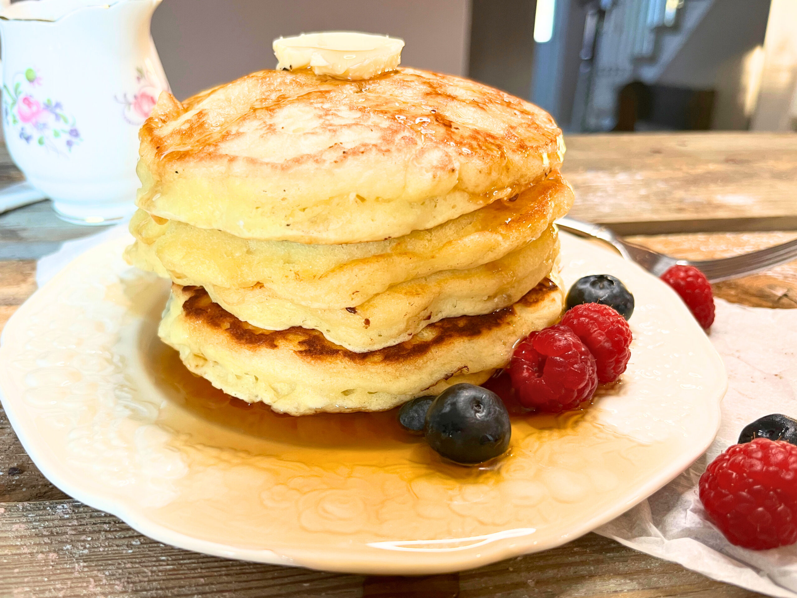 A stack of pancakes on a white plate. There are raspberries and blueberries around it. There is butter and maple syrup on top of the pancakes.
