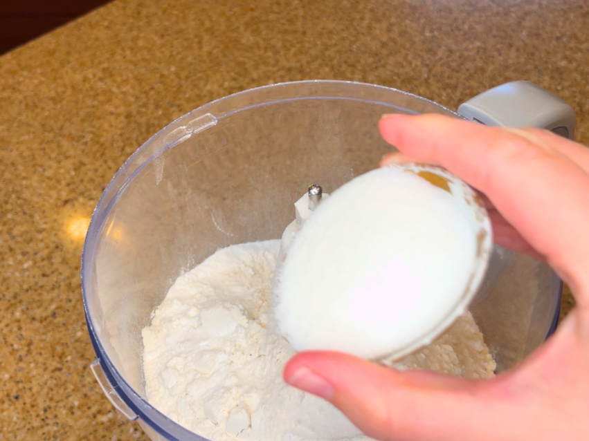 Woman pouring salt and baking powder in to a food processor with flour in it.