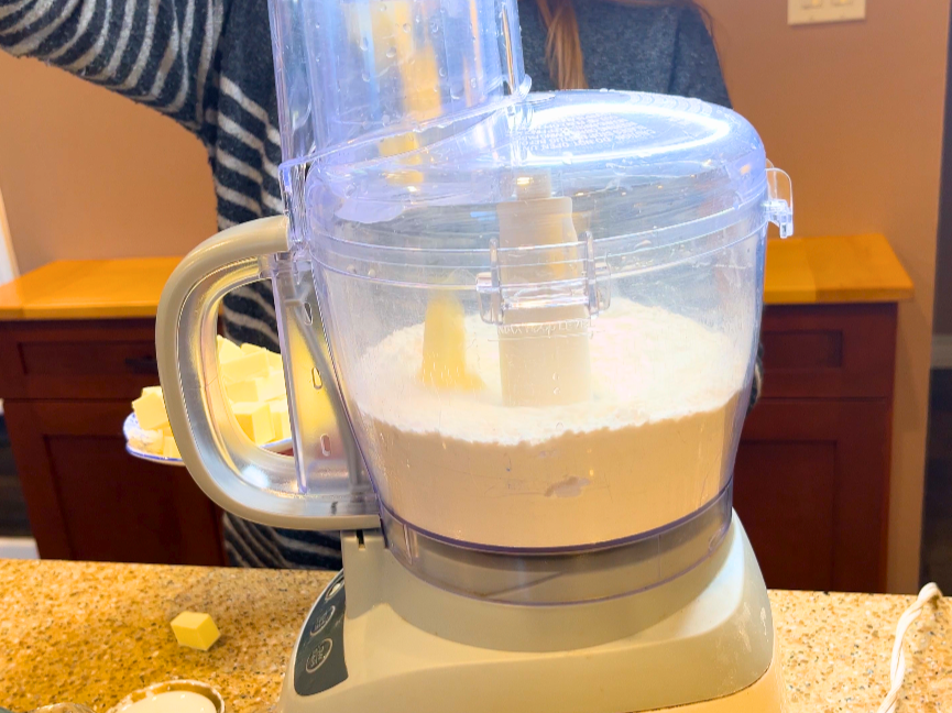 Woman adding cups of butter to a food processor with flour.