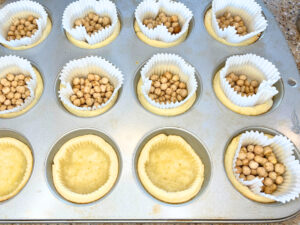 A muffin tin with liners and dried chickpeas inside. There are some with the liners removed to reveal partially cooked pie dough.