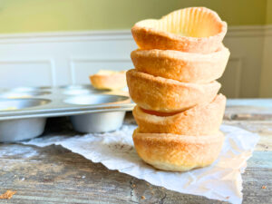 A stack of baked tart shells on a wooden table with a muffin tin in the background.