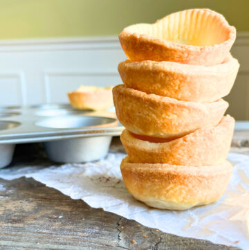 A stack of baked tart shells on a wooden table with a muffin tin in the background.