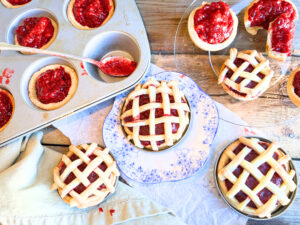 A wooden table with raspberry tarts on it. Some have a lattice top and some have an open top. One is on a blue floral plate and the background has tarts in a muffin tin.