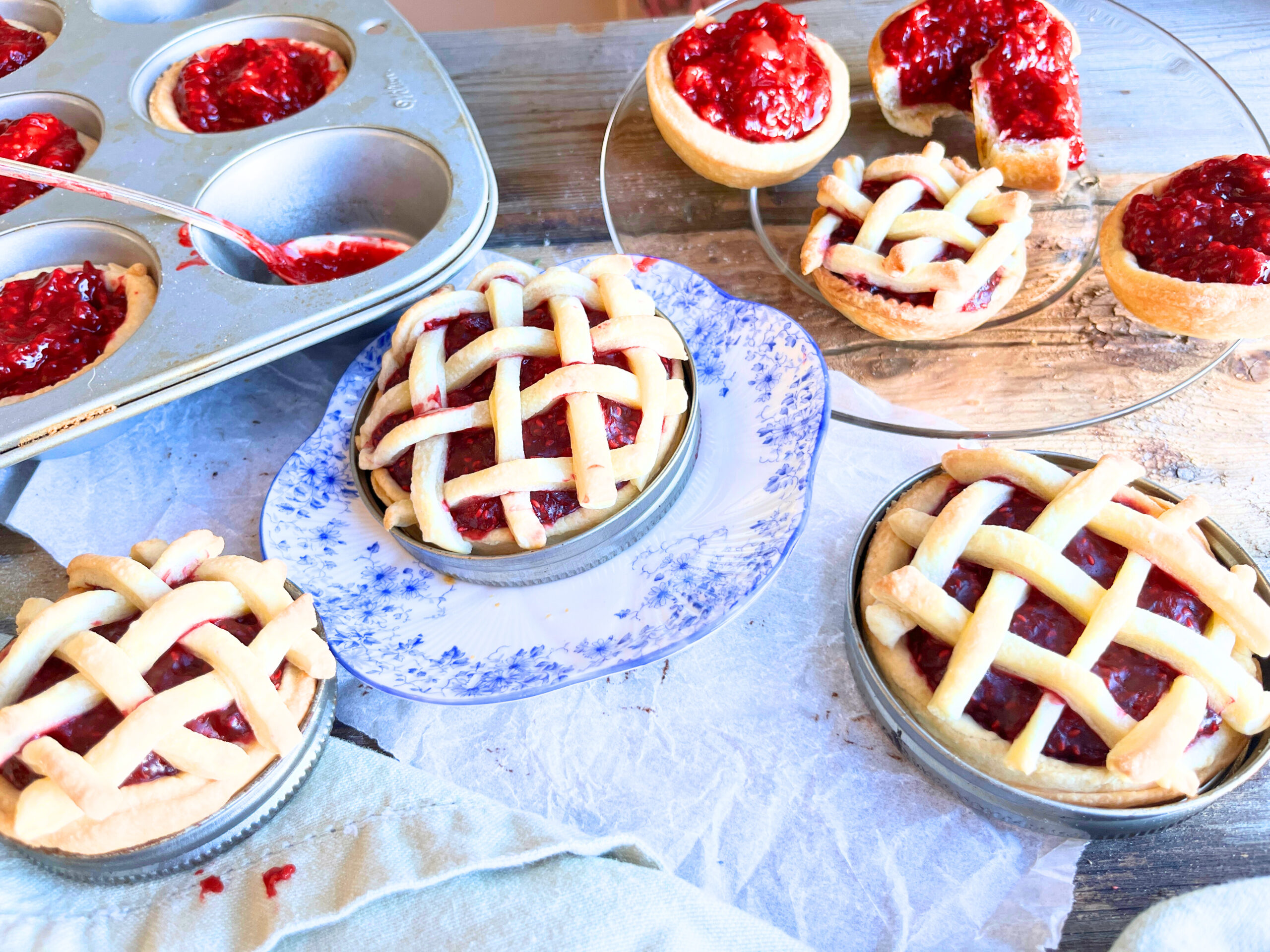 A wooden table with raspberry tarts on it. There are some with a lattice top and some with an open top. There is a muffin tray in the background.
