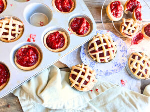 A wooden table with raspberry tarts on it. Some are in a muffin tin. Some have a lattice top and some have an open top.