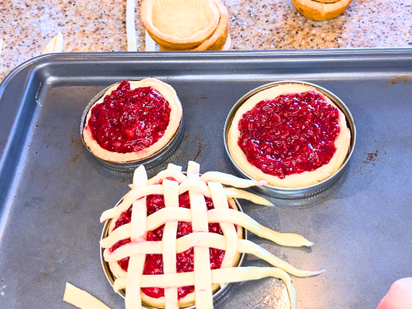 Three tart shells in mason jar lids. They raspberry filling and one has a lattice top being formed.