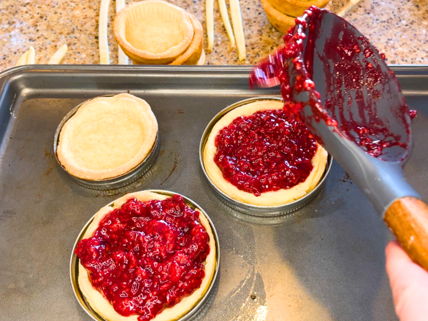 A woman spooning raspberry pie filling into baked tart shells.