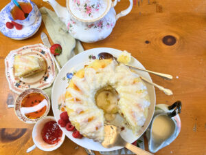 A vanilla cake with a white icing glaze sitting on a cake pedestal. There are teacups and a tea pot on the background. There is a slice out fo the cake.