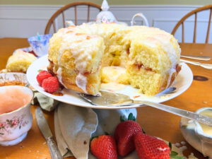 A vanilla cake with red fruit filling. There is a white icing glaze on top of the cake. There are teacups and a teapot in the background.