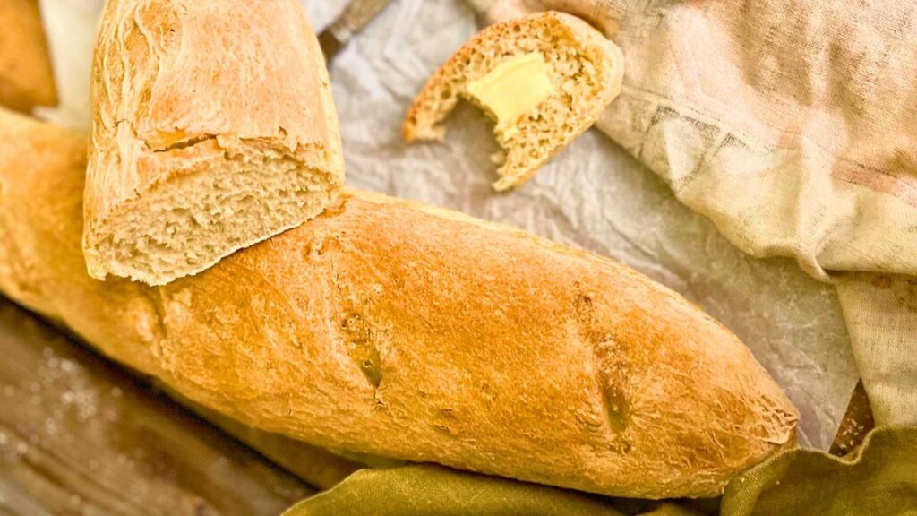 A baguette on a wooden table. There is a baguette on top that has been sliced.