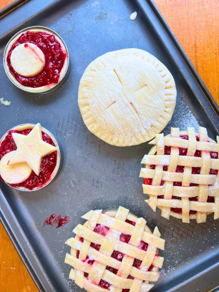 A baking sheet with Mason jar lid raspberry tarts. Two have a lattice top, one is a full pie crust, and 2 have cut-out tops.