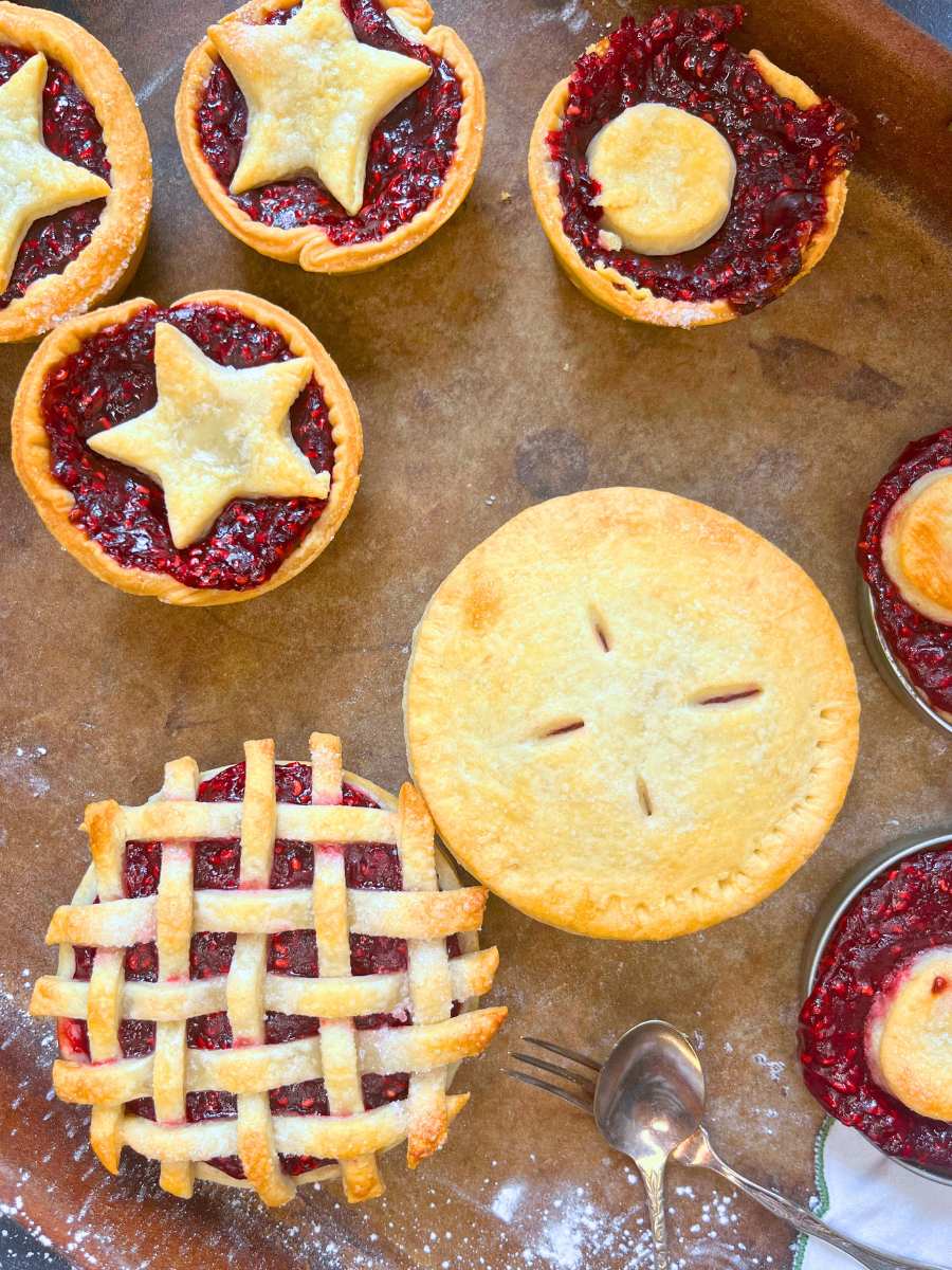 Assorted raspberry mini tarts on a stone baking sheet. Some have pastry cut-out tops, some lattice tops, and some full pastry tops.