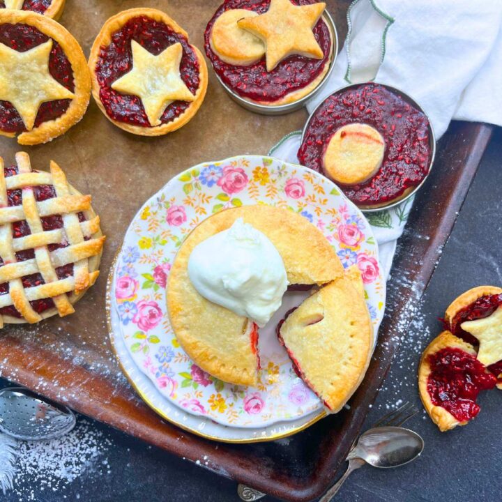 A stone baking sheet with mini raspberry tarts. Some have lattice tops some have pasty cut-out tops and some have full double pie crust. One is cut and has whipped cream on top.