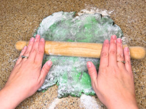 A woman rolling out green and black cookie dough with a wooden rolling pin.