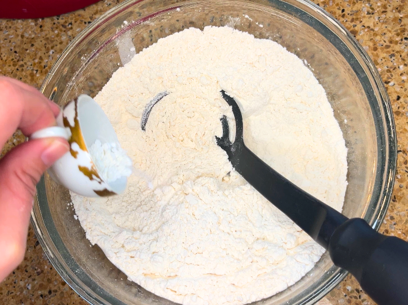 A woman pouring baking powder from a small cup into a large glass bowl with flour in it.