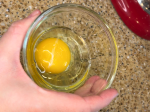 A woman holding a small glass bowl with a cracked egg inside.