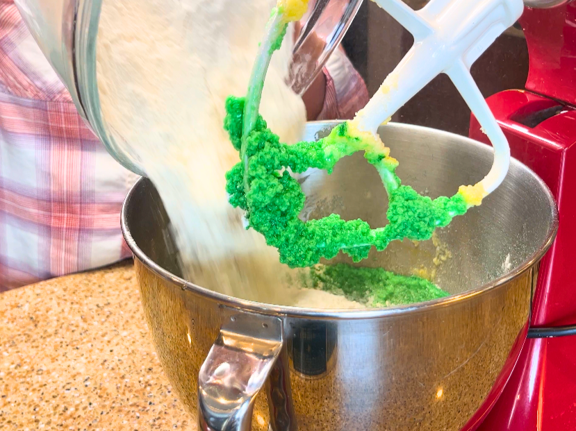 A woman adding flour to a stand mixer with a green batter inside.