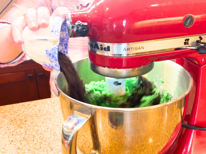A woman adding Oreo crumbles to a stand mixer with a green cookie dough inside.