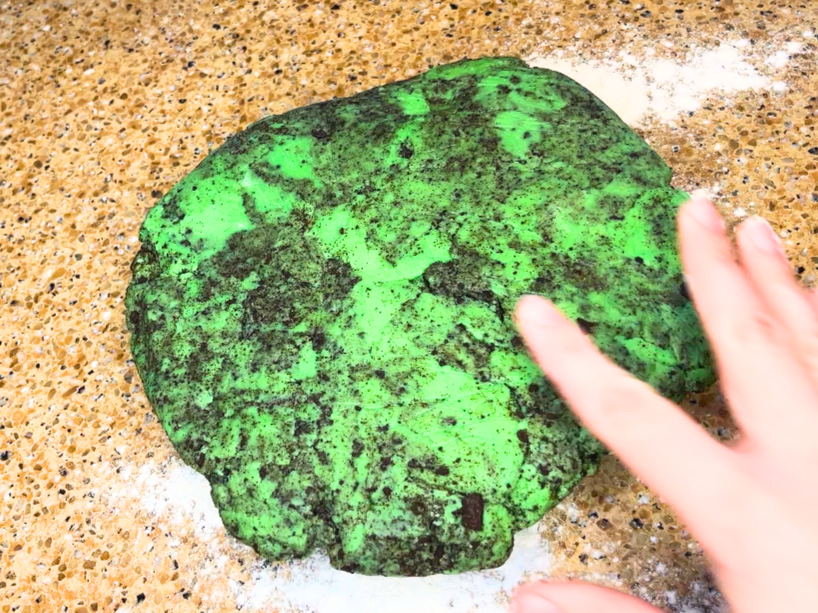 A woman placing green and black cookie dough on a floured counter top.