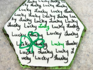 A sugar cookie decorated with royal icing. It has written words on it and a piped clover design.