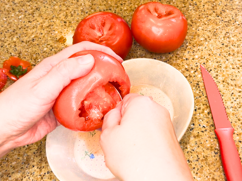 A woman scooping the pulp out of a tomato with a spoon.
